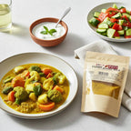 Curry Powder Mild pouch beside a bowl of vegetable curry, yogurt sauce, and a cucumber-tomato salad.