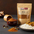 Garam masala pouch beside a bowl and pile of ground spice, whole spices, rice, and curry on a dark surface.