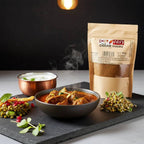 Garam masala pouch behind a bowl of curry with steam rising, with yogurt and lentil salads on a slate board.