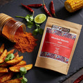 Mumbai Masala mix pouch on a dark surface with spilled spice from a jar, red chillies, lime, coriander, and potato wedges.